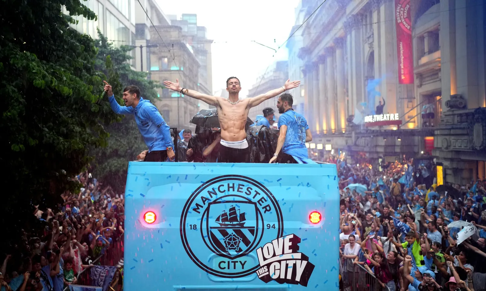 Manchester City players celebrating on the back of an open-top bus in the rain as fans line a Manchester street